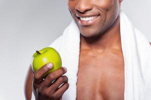Mann mit grünem Apfel. Zugeschnittenes Bild eines jungen muskulösen Afrikaners mit Handtuch auf der Schulter, der einen Apfel hochwirft und in die Kamera lächelt, während er vor grauem Hintergrund steht foto