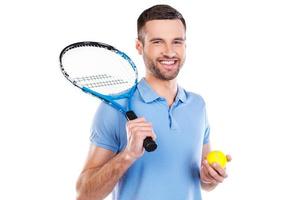bereit für ein großes Spiel. überzeugter junger Mann, der Tennisschläger hält und bei der Stellung gegen weißen Hintergrund lächelt foto