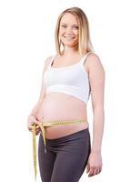 Messen ihres schwangeren Bauches. Schöne schwangere Frau, die ihren schwangeren Bauch misst und lächelt, während sie isoliert auf Weiß steht foto