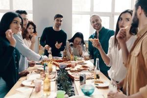 Pizza Party. Gruppe junger Leute in Freizeitkleidung, die Pizza essen und lächeln, während sie drinnen eine Dinnerparty haben foto