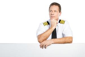 nachdenklicher Pilot. Nachdenklicher männlicher Pilot in Uniform, der sich an den Kopierraum lehnt und wegschaut, während er vor weißem Hintergrund steht foto