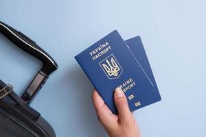Pässe eines Bürgers der Ukraine in weiblicher Hand auf blauem Hintergrund mit Koffer, Nahaufnahme. Auswanderungskonzept. Inschrift im ukrainischen ukrainischen Pass foto