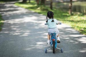 Rückseitenansicht des asiatischen Mädchenkindes, das lernt, Fahrrad im Parkgarten zu fahren. bildungskonzept für kinder üben radfahren im park, babysportkonzept. foto