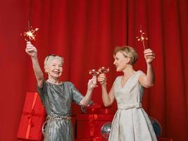 Zwei wunderschöne, stilvolle, reife Seniorinnen in Kleidern mit Wunderkerzen und Gläsern, die das neue Jahr feiern. weihnachten, familie, freunde, neujahr, spaß, party, stil, feierkonzept foto