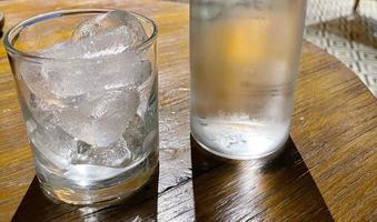 Selektiver Fokus, ein mit Eis gefülltes klares Glas und eine klare Glasflasche hält kaltes Mineralwasser auf einem Holztisch foto