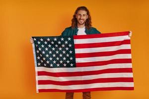 Fröhlicher junger Mann in Freizeitkleidung, der in die Kamera schaut und lächelt, während er die amerikanische Flagge vor gelbem Hintergrund hält foto