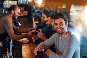 lächelnde junge Männer in Freizeitkleidung, die Bier trinken und sich zusammenschließen, während sie in der Kneipe sitzen foto