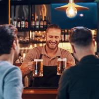 Fröhlicher Barkeeper, der jungen Männern Bier serviert, während er an der Bartheke im Pub steht foto