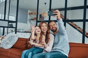 Verspielte junge Familie lächelt und macht Selfie per Smartphone, während sie Zeit zu Hause verbringt foto