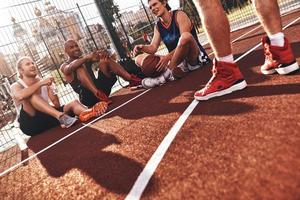 Zeit zusammen verbringen. Gruppe junger Männer in Sportkleidung lächelnd, während sie draußen auf dem Basketballfeld sitzen foto