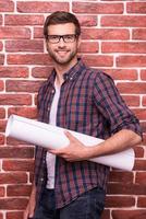 selbstbewusster Architekt. hübscher junger Mann mit Brille, der eine Blaupause hält und lächelt, während er gegen eine Ziegelwand steht foto