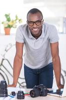 junger und kreativer Arbeiter. Fröhlicher junger afrikanischer Mann in Freizeitkleidung lehnt am Tisch mit Kamera und Objektiv darauf foto