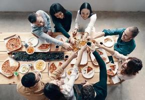 Gruppe junger Leute in Freizeitkleidung, die Pizza pflücken und lächeln, während sie drinnen eine Dinnerparty haben foto