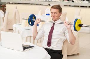 sportlicher Geschäftsmann. lächelnder junger Mann mit roten Haaren in Hemd und Krawatte, der im Restaurant sitzt und eine Hantel hält foto