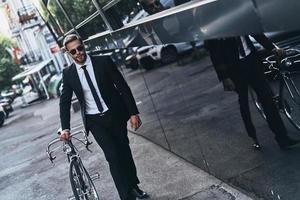 umweltfreundlich. hübscher junger mann im vollen anzug, der sein fahrrad zieht, während er im freien geht foto