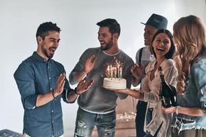 Überraschen Sie den glücklichen jungen Mann, der gestikuliert und lächelt, während er unter Freunden Geburtstag feiert foto