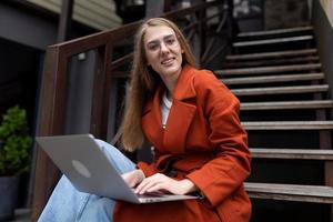 junge Frau, die online auf einem Laptop in einem Herbstmantel arbeitet und auf der Treppe in der Stadt sitzt foto