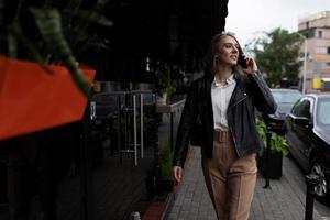 Stilvolle junge Frau, die die Straße hinuntergeht und mit einem Lächeln telefoniert und sich umschaut foto