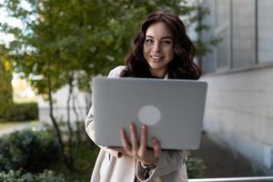 Stilvolle junge Geschäftsfrau mit Laptop in den Händen vor dem Hintergrund eines Bürogebäudes, starkes und unabhängiges Frauenkonzept foto