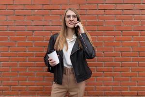 Junge Studentin, die auf einem Handy mit einer Tasse Kaffee in den Händen vor dem Hintergrund einer orangefarbenen Backsteinmauer spricht foto