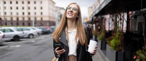 Junge starke Frau, die mit einer Tasse Kaffee in den Händen durch die Stadt läuft, blickt verträumt in die Ferne foto