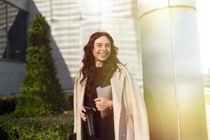 Stilvolle Unternehmerin mit einem breiten Lächeln auf dem Hintergrund eines Bürogebäudes mit einem Laptop in den Händen, Karrierewachstumskonzept foto