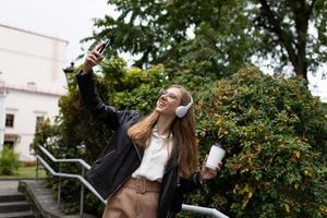 Junge glückliche Frau, die per Videoanruf in Overhead-Kopfhörern mit einer Tasse Kaffee in den Händen spricht und vor dem Hintergrund eines Stadtparks tanzt foto