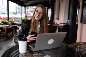 junge freiberufliche frau, die kaffee trinkt und online auf einem laptop in einem stadtcafé arbeitet, mit einem lächeln, das in die kamera schaut foto
