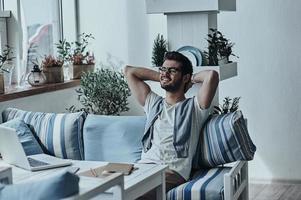 Freude an den Ergebnissen seiner Arbeit. hübscher junger moderner Mann in Brillen, der die Hände hinter dem Kopf hält und lächelt, während er auf dem Sofa im Restaurant sitzt foto