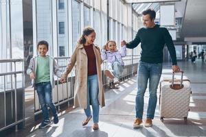 volle Länge der glücklichen Familie mit zwei kleinen Kindern, die am Flughafenterminal spazieren gehen foto