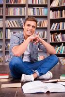 Zeit in der Bibliothek verbringen. glücklicher junger mann, der hände am kinn hält und lächelt, während er gegen bücherregal sitzt foto