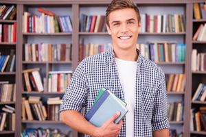 Studentin in der Bibliothek. hübscher junger Mann, der Bücher hält und lächelt, während er in der Bibliothek steht foto