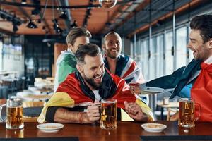 Fröhliche junge Männer, die mit internationalen Flaggen bedeckt sind, Bier trinken und sich zusammenschließen, während sie in der Kneipe sitzen foto