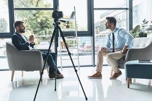 Interview filmen. Zwei junge Männer in eleganter Freizeitkleidung unterhalten sich, während sie drinnen ein neues Video drehen foto