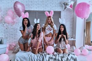 Mädchenzeit. Vier verspielte junge Frauen in Hasenohren, die ein Gesicht machen und lächeln, während sie auf dem Bett sitzen foto