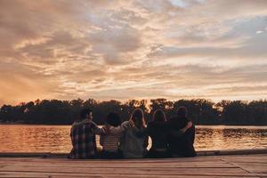 Leben voller Freundschaft. Rückansicht junger Menschen in Freizeitkleidung, die sich umarmen, während sie auf dem Pier sitzen foto