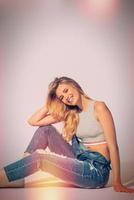 unbeschwert und schön. Studioaufnahme einer schönen jungen Frau in Jeans, die insgesamt in die Kamera schaut und lächelt, während sie auf dem Boden sitzt foto