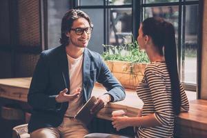 Geschäft besprechen. Fröhlicher junger Mann und Frau, die etwas diskutieren und lächeln, während sie beide an der Holztheke im Café sitzen foto