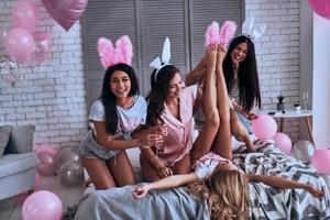machen was sie wollen. Verspielte junge Frauen, die versuchen, Hasenohren an den Füßen ihrer Freundin anzuziehen und lächeln, während sie auf dem Bett sitzen foto