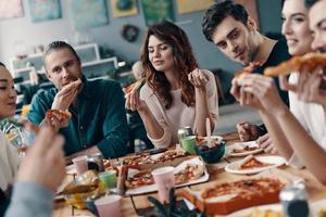 leckeres essen teilen. Gruppe junger Leute in Freizeitkleidung, die Pizza essen und lächeln, während sie drinnen eine Dinnerparty haben foto