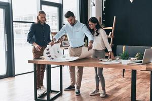 Strategie besprechen. in voller Länge von den jungen selbstbewussten Geschäftsleuten, die etwas besprechen, während sie Blaupause im Büro betrachten foto