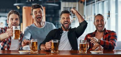 Jubelnde junge Männer in Freizeitkleidung, die Bier trinken und Sportspiele beobachten foto