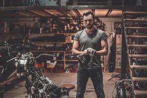 überzeugter Mechaniker. Selbstbewusster junger Mann mit Lappen und Blick in die Kamera, während er in der Werkstatt neben dem Motorrad steht foto