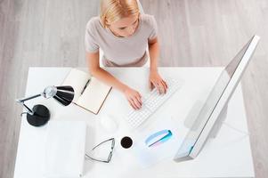 Geschäft zuerst. Blick von oben auf die schöne junge Frau mit blonden Haaren, die am Computer arbeitet, während sie an ihrem Arbeitsplatz sitzt foto