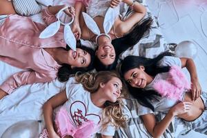 extrem schön. Draufsicht von vier attraktiven jungen Frauen, die lächeln, während sie zu Hause auf dem Bett liegen foto