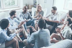 applaudieren ihren Leistungen. Gruppe junger, fröhlicher Menschen, die im Kreis sitzen und applaudieren, während ein Mann gestikuliert und lächelt foto