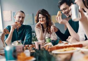 lassen Sie alle Sorgen hinter sich. Gruppe junger Leute in Freizeitkleidung, die Pizza essen und lächeln, während sie drinnen eine Dinnerparty haben foto