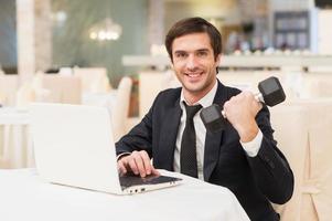 Sport und Wirtschaft. lächelnder junger Mann in Abendkleidung, der am Laptop sitzt und eine Hantel hält foto