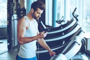 beste Lied für sein Training. Seitenansicht eines jungen gutaussehenden Mannes in Sportbekleidung, der sein Smartphone benutzt, während er im Fitnessstudio auf dem Laufband steht foto