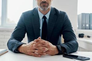Nahaufnahme eines Mannes in formeller Kleidung, der die Hände gefaltet hält, während er am Schreibtisch im Büro sitzt foto